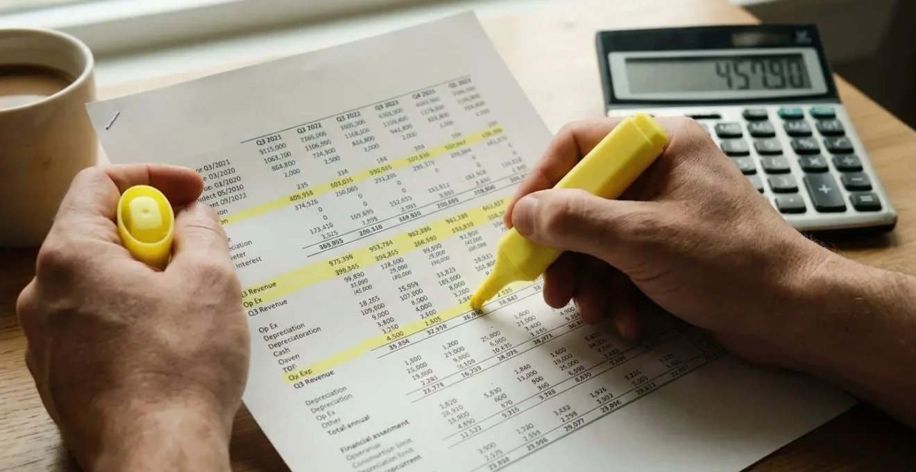 Gros plan sur des mains tenant un surligneur jaune annotant un document financier imprimé, calculatrice floue en arrière-plan